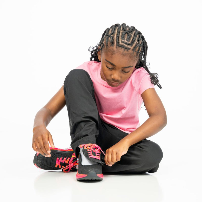 A young girl with braided hair in a pink shirt and black pants sits on a white floor, focused as she ties the laces of her BILLY Footwear SALE - Black/Fuchsia Fire BILLY Sport Inclusion shoes.