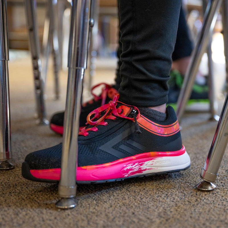 Close-up of a person in SALE - Black/Fuchsia Fire BILLY Sport Inclusion shoes by BILLY Footwear, featuring bright pink and orange accents, standing on carpet among metal chair legs.