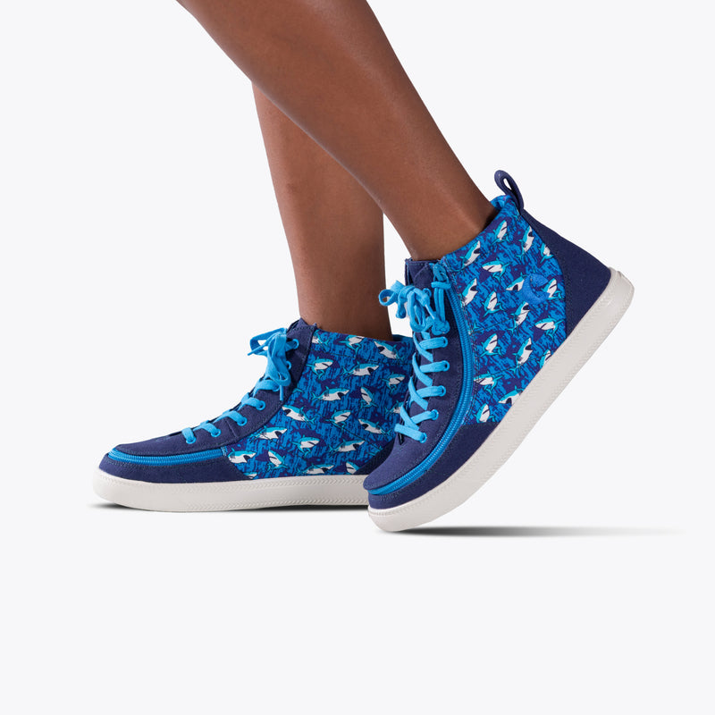 A child sports the SALE - Blue Sharks BILLY Classic Lace High from BILLY Footwear—kids’ high-top sneakers with blue shark patterns, blue laces, white soles, and a wraparound zipper, set against a plain white background.