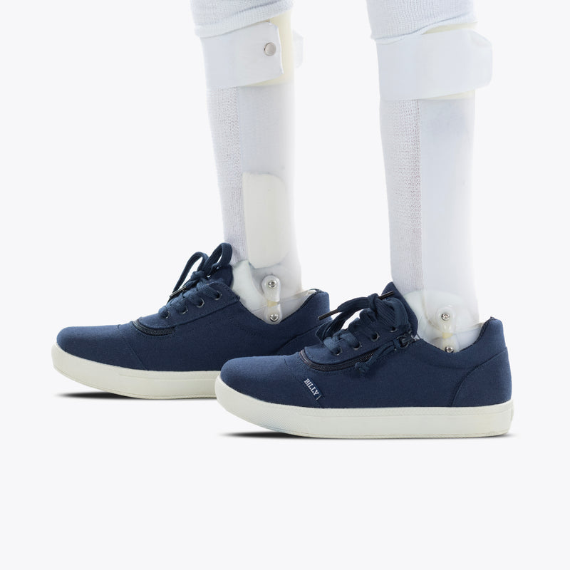 A person wearing the SALE - Navy BILLY D|R II Short Wrap Low shoes by BILLY Footwear, featuring white soles and compatible with AFOs and prosthetic legs, stands on a white surface.