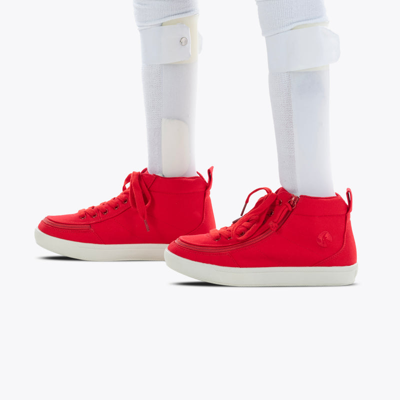A person with visible white prosthetic legs wears the SALE - Red BILLY Classic D|R II High by BILLY Footwear. These red high-top, AFO-friendly shoes feature laces, side zippers, and white soles, shown on a plain white background.