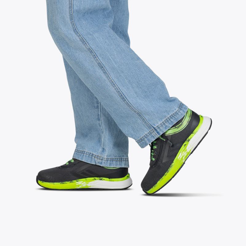 A person wears blue jeans and black SALE - Smoke/Lime Fire BILLY Sport Inclusion shoes by BILLY Footwear, featuring neon green accents, standing with one foot ahead against a white background to showcase the stylish athletic zippered design.