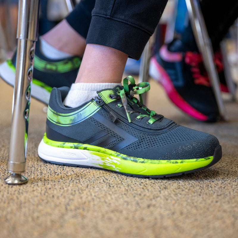 Someone wearing SALE - Smoke/Lime Fire BILLY Sport Inclusion sneakers by BILLY Footwear with white socks sits on a tan carpeted floor, one foot tucked under their chair. In the background, another person wears black and pink shoes.