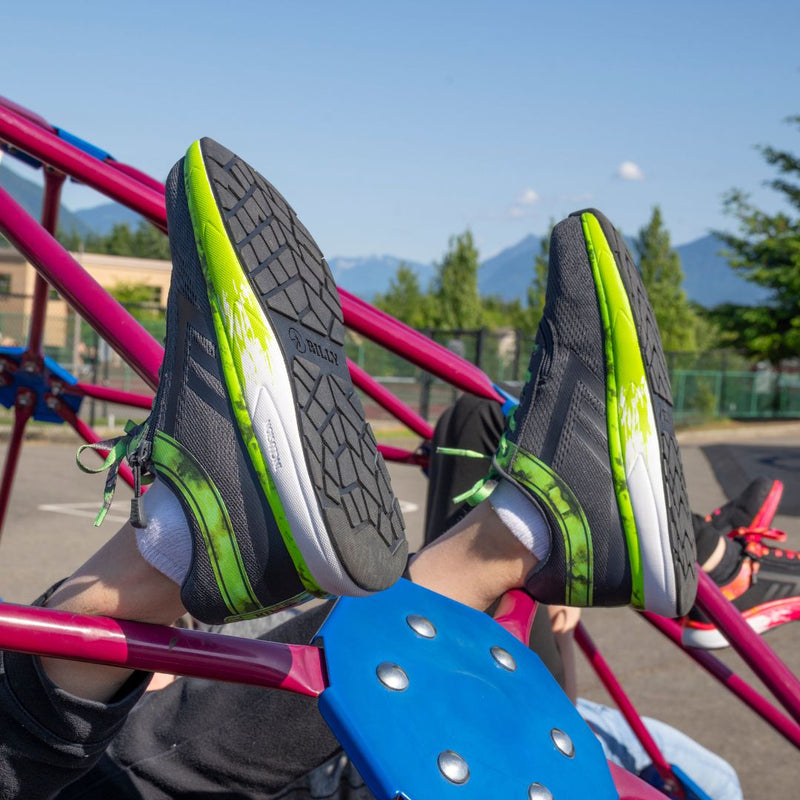 A person wearing SALE - Smoke/Lime Fire BILLY Sport Inclusion shoes from BILLY Footwear with white socks is lying on playground equipment, soles visible. Other people and mountains are in the background.