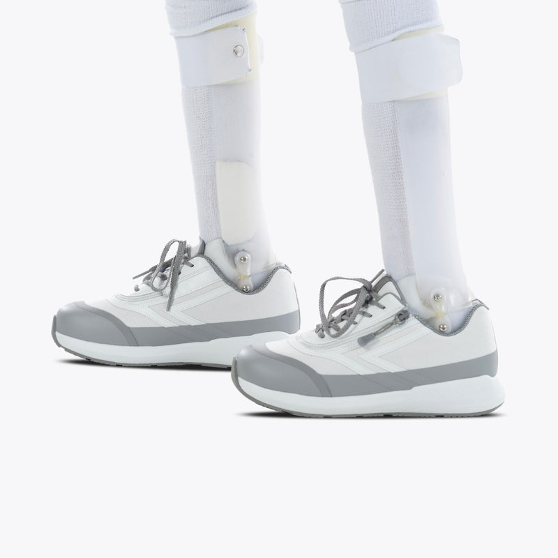Close-up of two prosthetic legs fitted with SALE - White BILLY Goat shoes by BILLY Footwear, featuring an easy on-and-off zipper, standing on a white background.