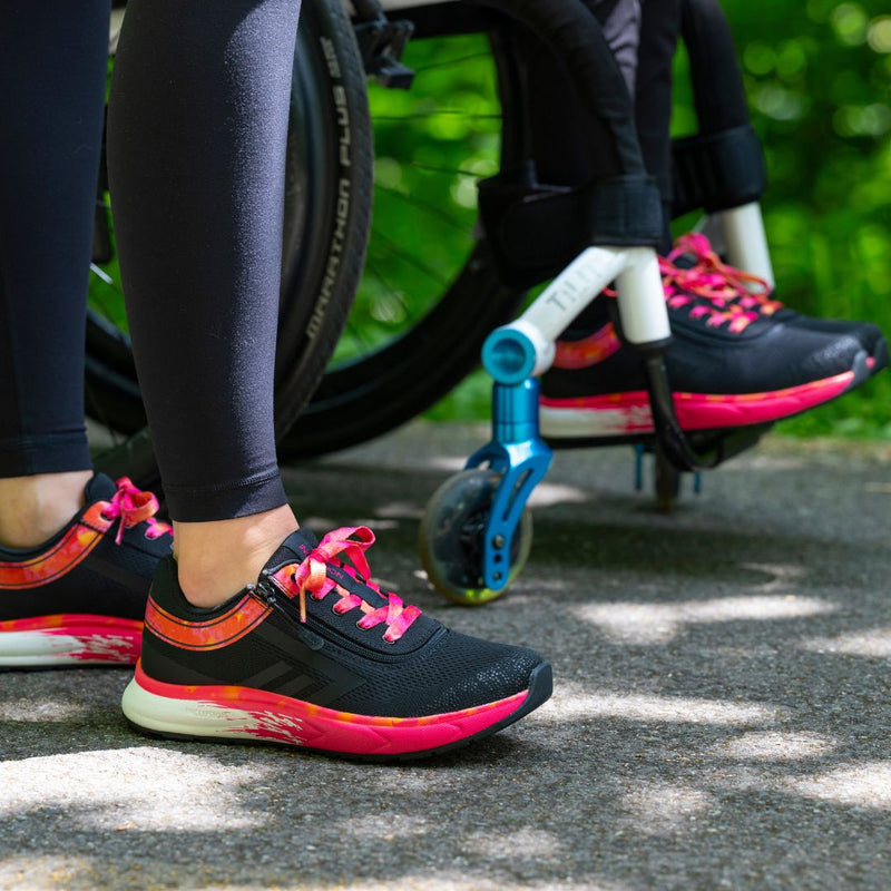A person in black leggings stands next to someone in a wheelchair outdoors, both wearing BILLY Footwear’s SALE - Women's Black/Fuchsia Fire BILLY Sport Inclusion shoes with cushioned insoles as sunlight filters through green foliage.