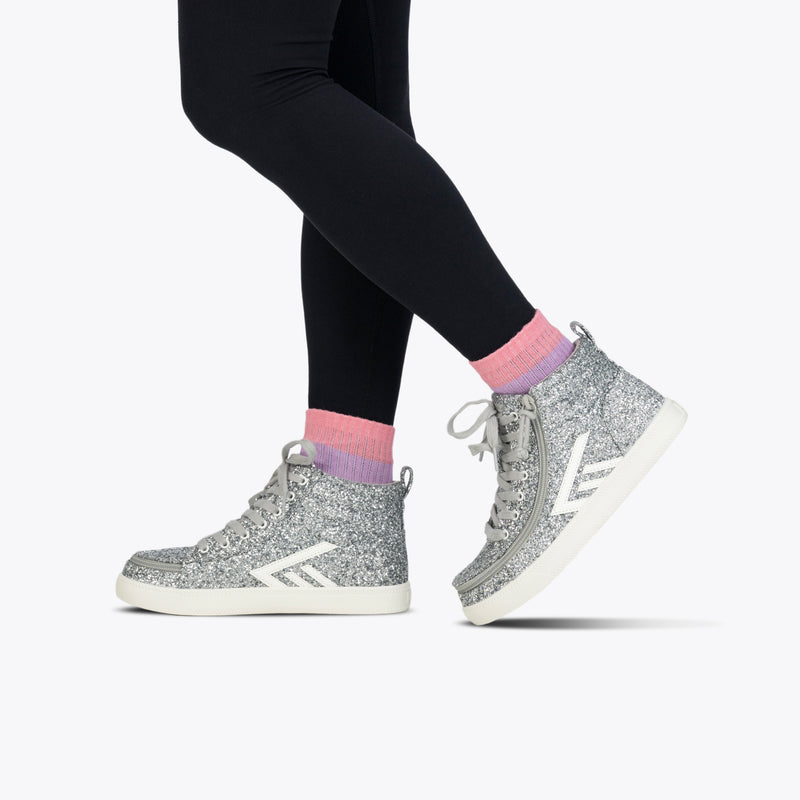 A person stands on tiptoes against a plain light background, wearing black leggings, pink and lavender socks, and BILLY Footwear's Silver Glitz BILLY CS High zippered high tops—sparkly sneakers perfect as kids skate shoes.
