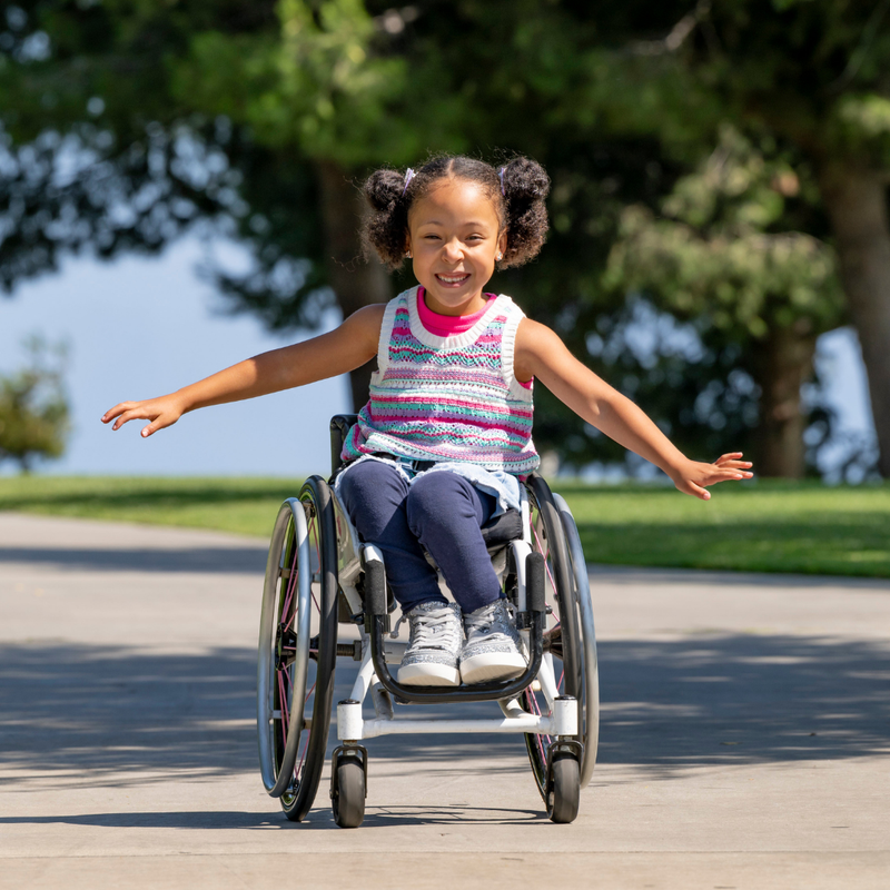 A young girl with pigtails smiles and spreads her arms while using a wheelchair outdoors on a sunny day, wearing Silver Glitz BILLY CS High sneakers by BILLY Footwear. Green grass and trees brighten the background.