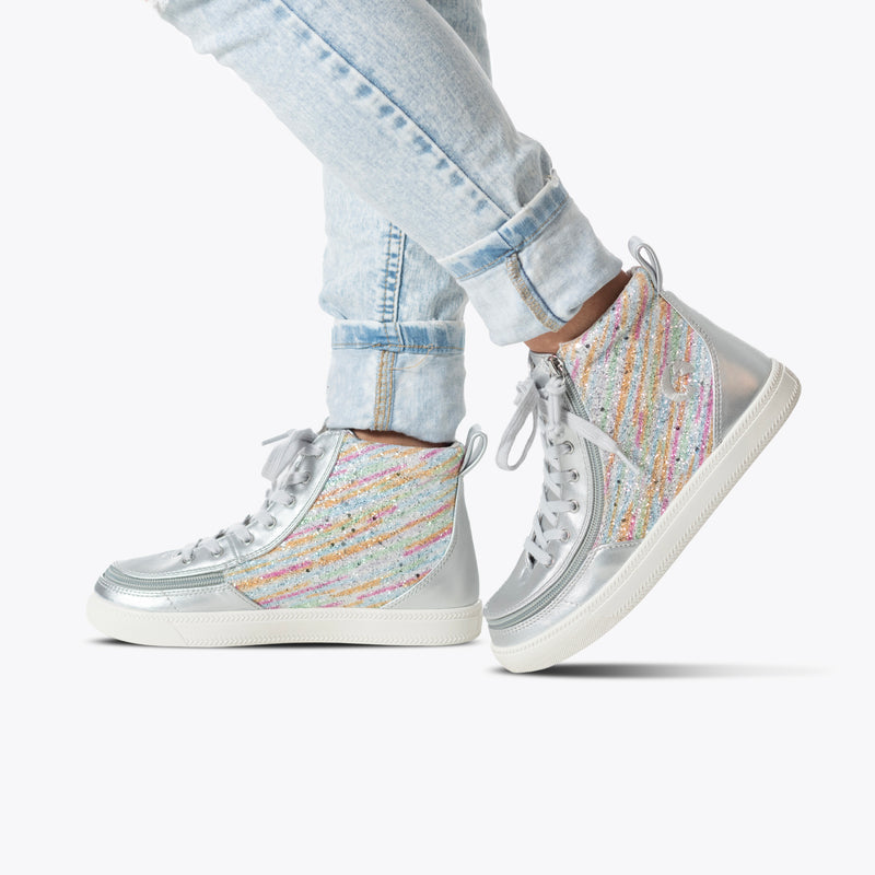 A person wears light-wash, cuffed jeans and the BILLY Footwear Silver Rainbow BILLY Classic Lace High kids sneakers—silver high tops with colorful glittery stripes and a wraparound zipper—photographed on a plain white background.