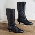 Pair of Veronica black knee-high boots with thin heels on a concrete surface.