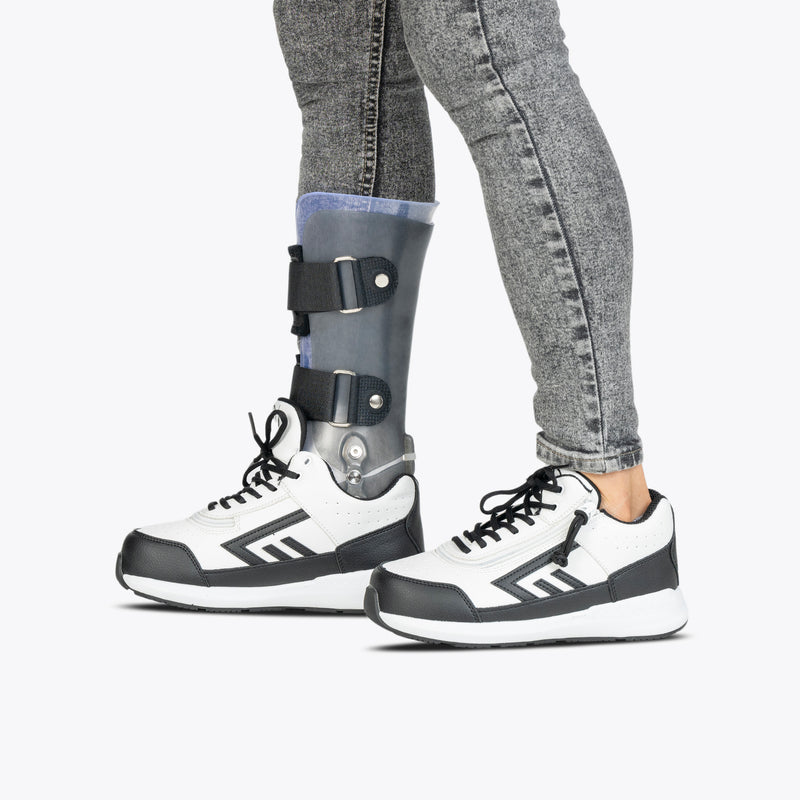A person in gray jeans and BILLY Footwear’s White/Black BILLY Goat Hoop sneakers, designed as orthotic shoes, stands with a prosthetic left leg against a plain white background.