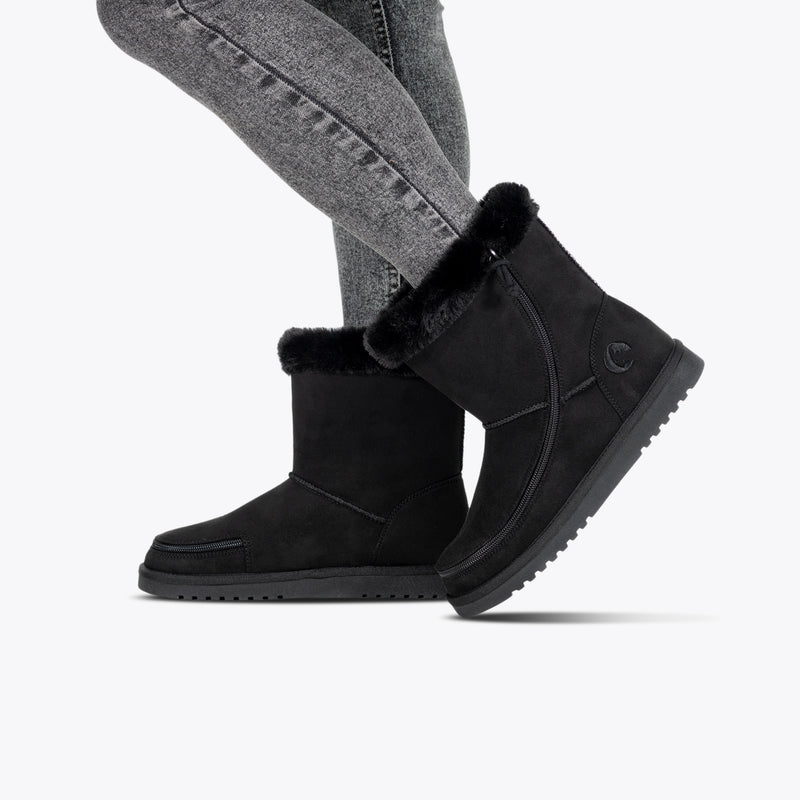 A person in gray jeans and BILLY Footwear Women's Black BILLY Cozy II zipper boots with flannel lining, round toe, and flat rubber sole walks against a plain light background.