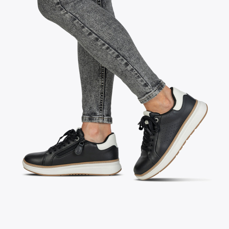 A person wears Women's Black Leather BILLY Comfort Luxe shoes by BILLY Footwear with white soles and gray skinny jeans, set against a plain, light background.