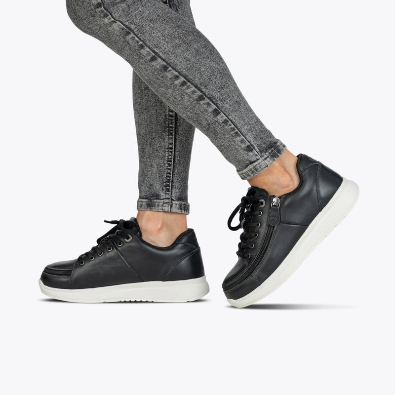 A person wears Women's Black Leather BILLY Comfort Plush shoes by BILLY Footwear with white soles and gray skinny jeans, posing with one foot slightly forward against a plain light background.