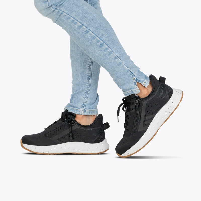 A person wearing light blue skinny jeans and Women's Black/White/Gum BILLY Sport Inclusion DZ1 sneakers by BILLY Footwear walks against a plain light background.