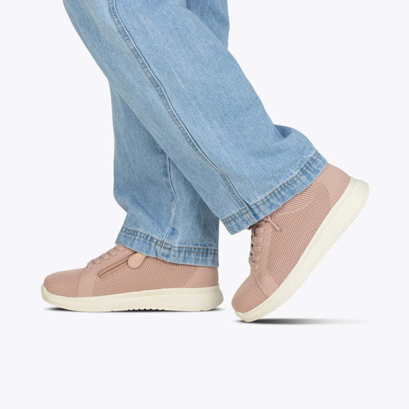 A person stands against a white background, one foot forward, wearing light blue jeans and Women's Blush BILLY Comfort Knit sneakers by BILLY Footwear, featuring breathable construction, white soles, and a medial zipper for comfort and style.