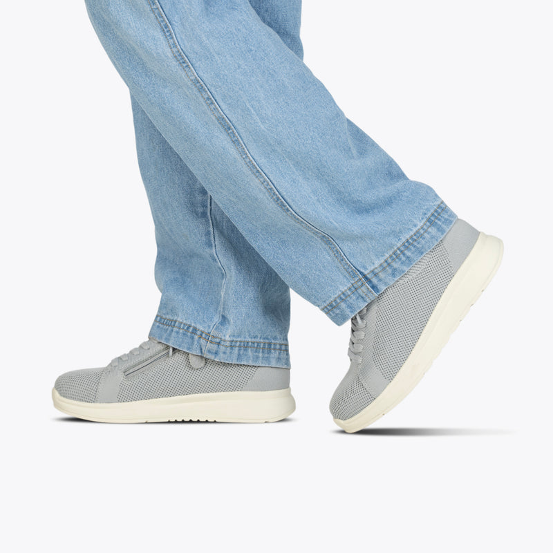 A person in light blue jeans and Women's Grey BILLY Comfort Knit sneakers by BILLY Footwear, featuring a breathable design and white soles, walks on a plain white background. Only the lower legs and feet are visible.