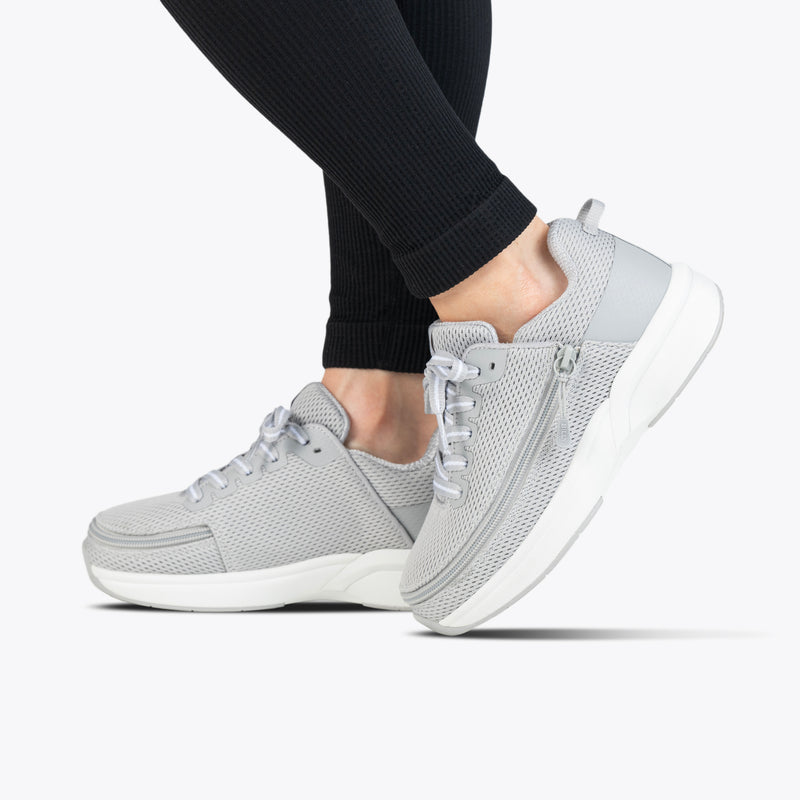 A person wearing black leggings and Women's Grey BILLY Limitless shoes by BILLY Footwear, shown from the side on a white background. The shoes have a light gray mesh upper, white soles, and a convenient side zipper detail.