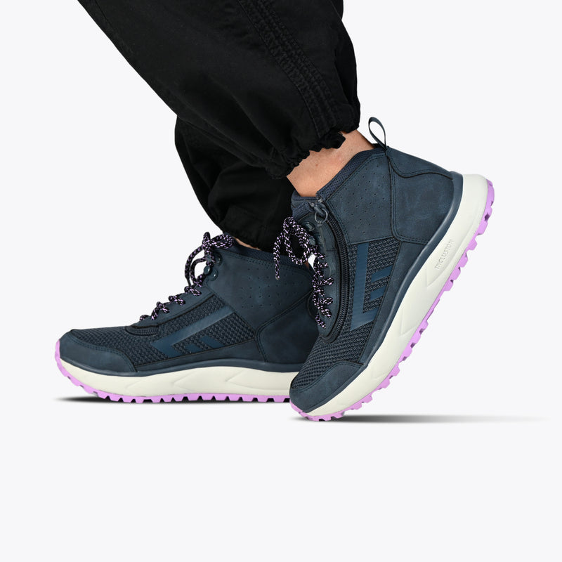 A person stands against a plain white background wearing black pants and BILLY Footwear Women's Navy/Purple BILLY Inclusion Trail Boots, featuring a high-top design, easy on/off zipper, white soles, and purple outsoles.