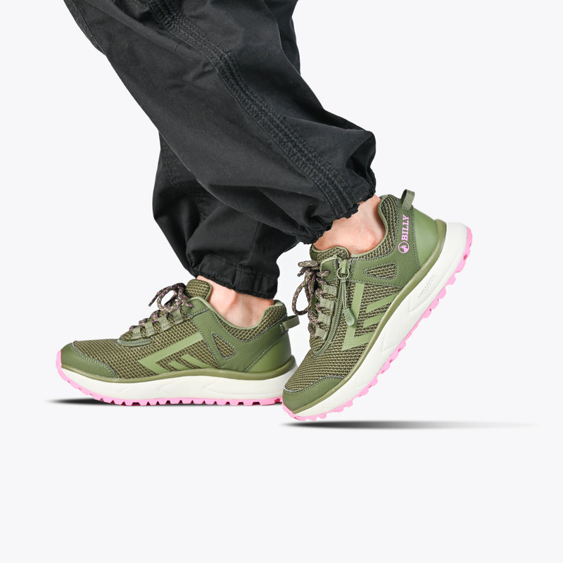 A woman stands on tiptoe in black pants and olive/pink BILLY Inclusion Trail sneakers by BILLY Footwear, featuring pink soles and a convenient easy on/off zipper, against a plain white background.