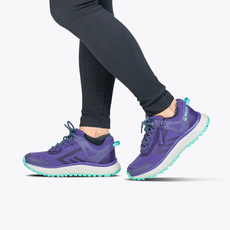 A person in black leggings and Women's Purple/Mint BILLY Inclusion Trail sneakers by BILLY Footwear, featuring an easy on-off zipper, walks against a plain white background.