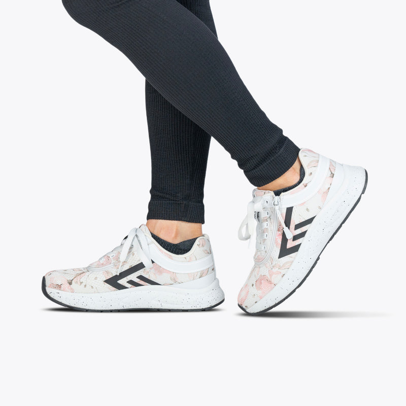 A person wearing black leggings and BILLY Footwear’s Women's Stone Floral BILLY Sport Inclusion athletic zipper shoes with floral prints and black chevron accents stands on tiptoe against a plain white background.