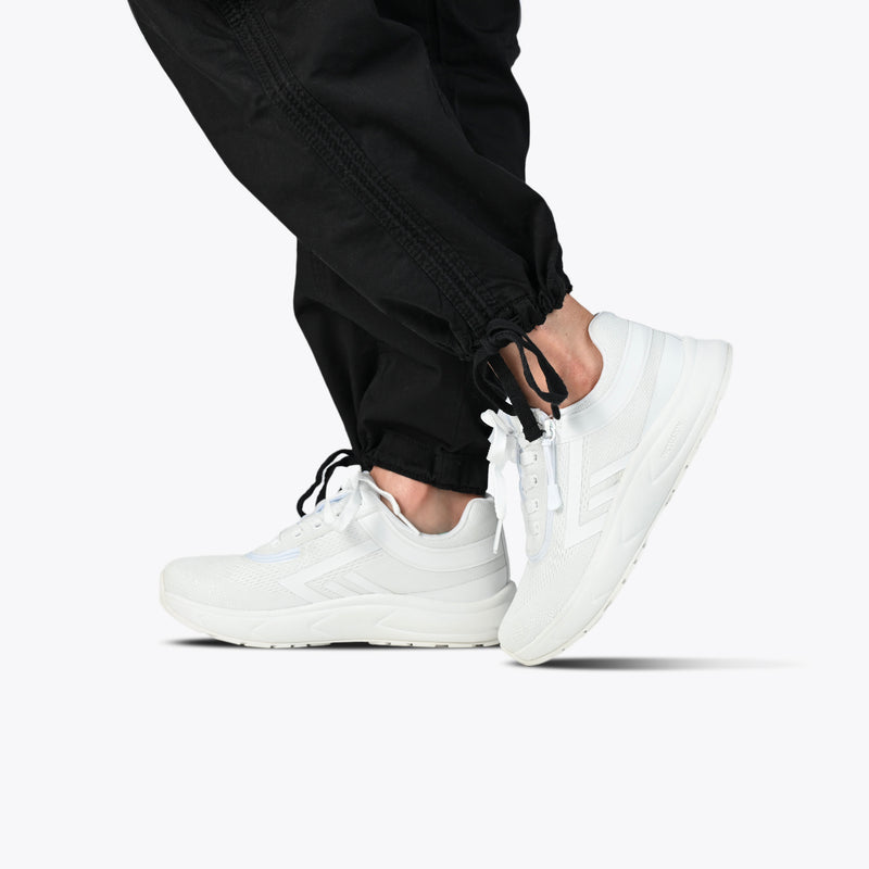 A person, shown from the knees down against a plain white background, walks wearing black jogger pants and Women's White BILLY Sport Inclusion shoes by BILLY Footwear.