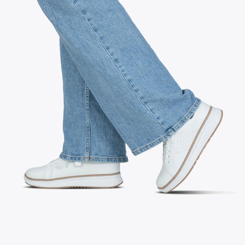 A person walks with one foot in front of the other, wearing light blue jeans and Women's White Leather BILLY Comfort Luxe shoes by BILLY Footwear, set against a plain white background.