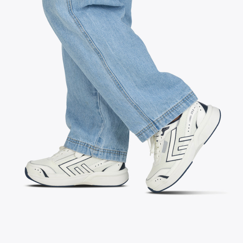 A person wearing light blue jeans and BILLY Footwear’s Women’s White/Navy BILLY Limitless 253 sneakers walks against a light background, with only their lower legs and feet visible.