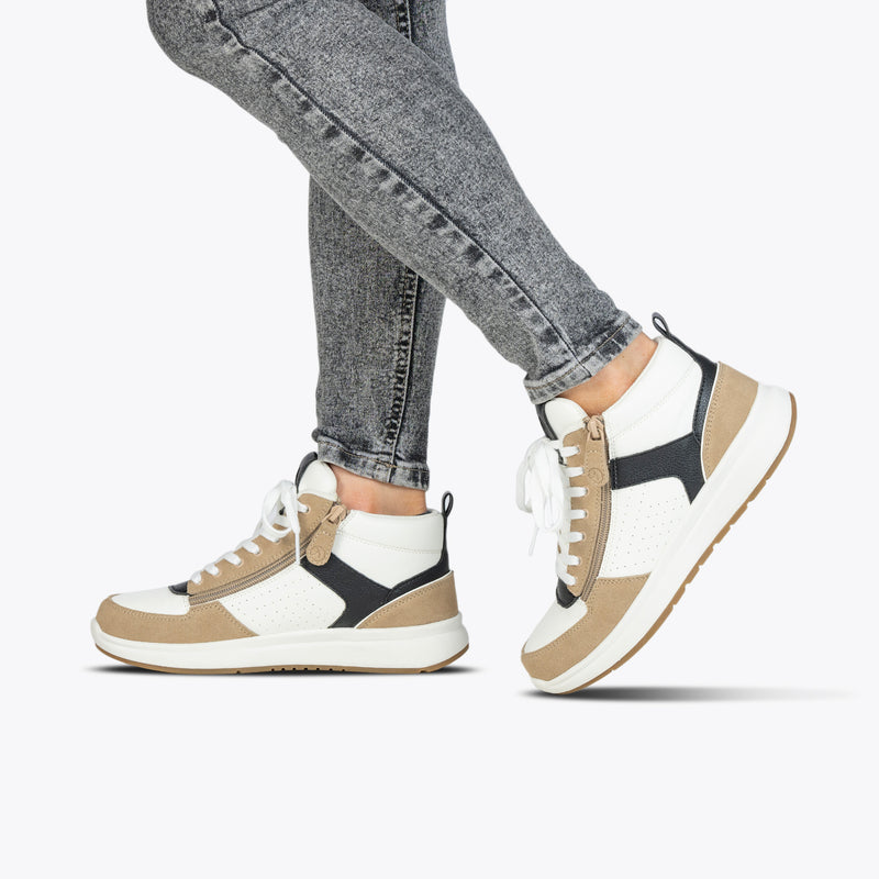 A person wearing grey jeans and BILLY Footwear Women's White/Tan/Black BILLY Comfort DZ High sneakers with double zippers stands on one foot, the other slightly raised, against a plain white background.