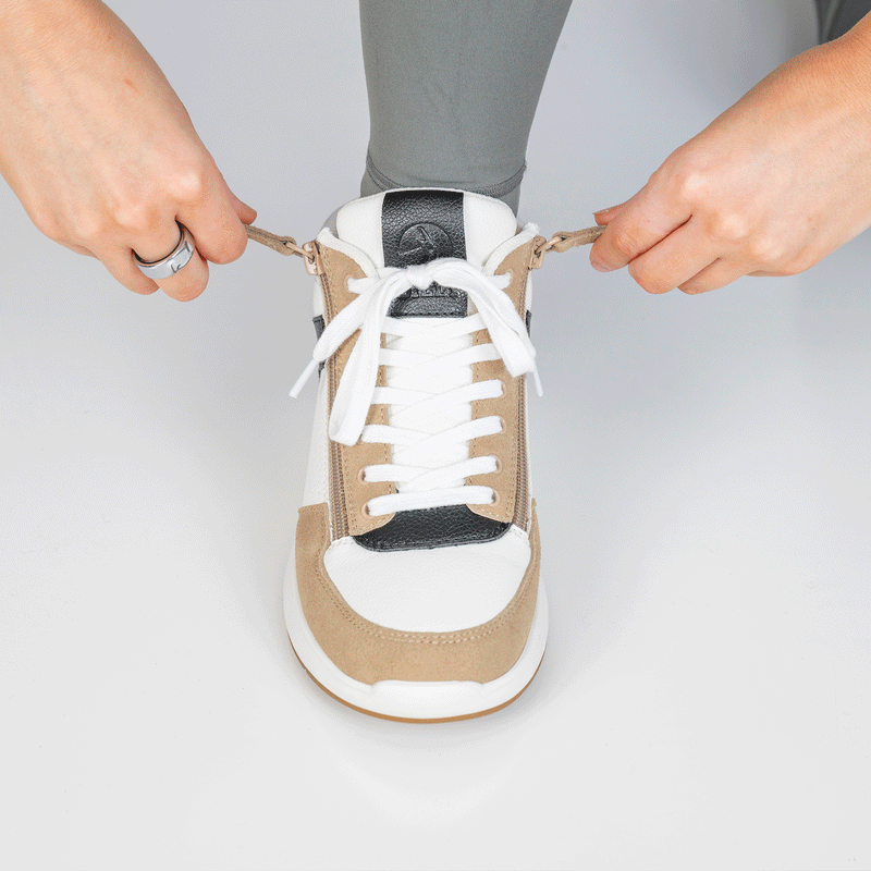 A person ties the laces of a Women's White/Tan/Black BILLY Comfort DZ High sneaker by BILLY Footwear, wearing gray leggings and a silver ring on their left hand, set against a plain white background.