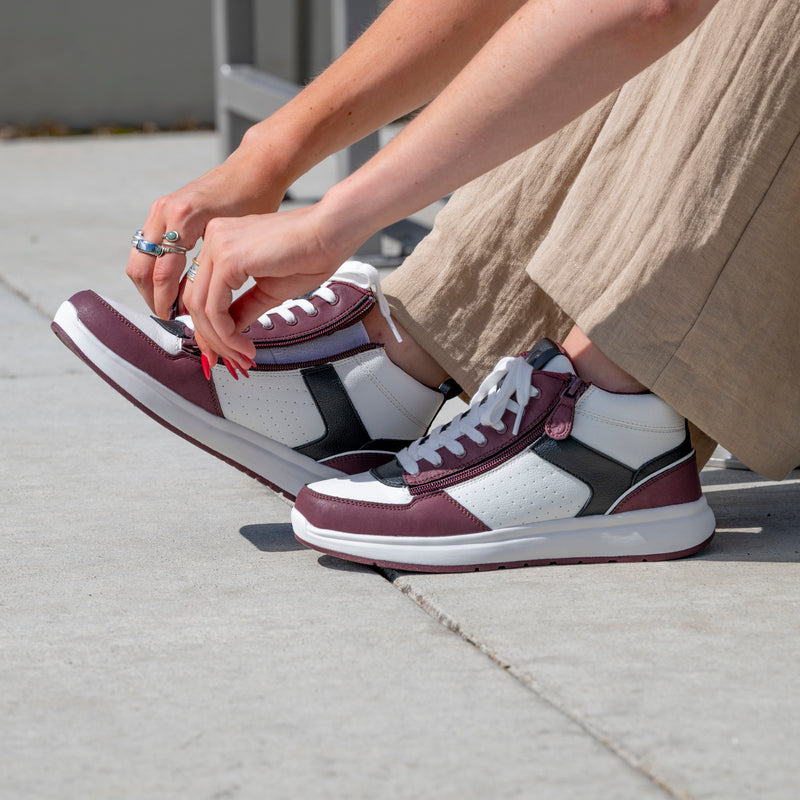 A woman in a beige skirt ties the laces of BILLY Footwear Women's White/Wine/Black BILLY Comfort DZ High sneakers while sitting outdoors on concrete.
